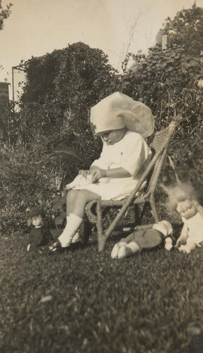 Black and white photograph of a young girl dressed in a nurse's outfit, seated on a chair in a garden. - click to view larger image