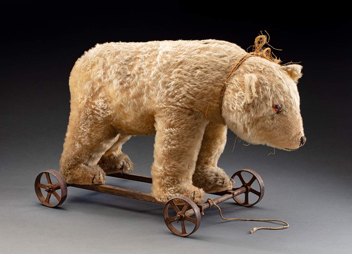 A stuffed pull along plush toy polar bear, standing on four paws, secured to a metal and wooden frame with three wheels. The fourth (rear left) wheel is missing. The polar bear has a pair of red glass or plastic eyes, and a black thread mouth, nose, and toes. There is a rope tied around its neck with another rope tied to the front of the metal frame. Some of the fur has fallen out, and the metal frame and the wheels are covered in rust. - click to view larger image