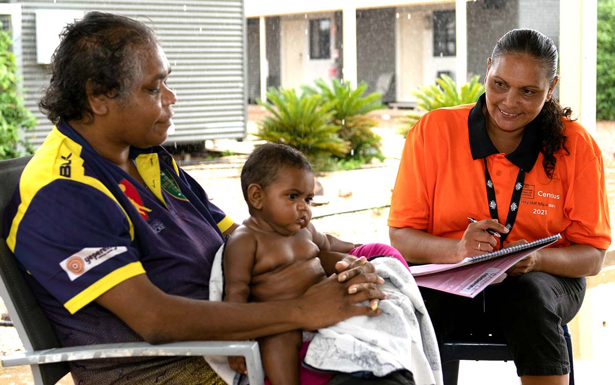 A woman is sitting in a chair with a child in her lap. A female Census staff member is smiling at the child with a pen and log book in her lap.