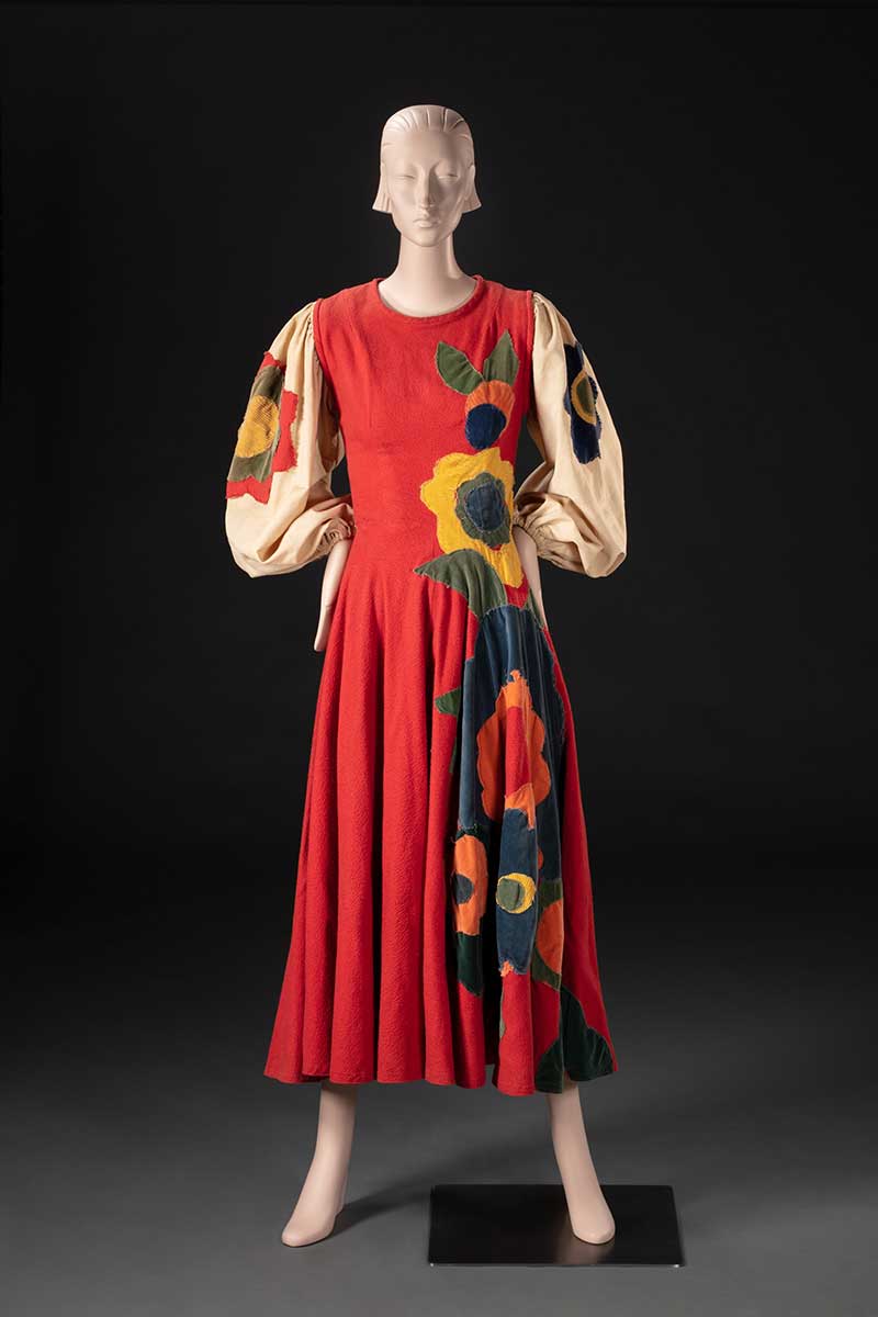 A mannequin displaying a costume featuring a full length red dress with loose beige sleeves, and floral embellishments. - click to view larger image