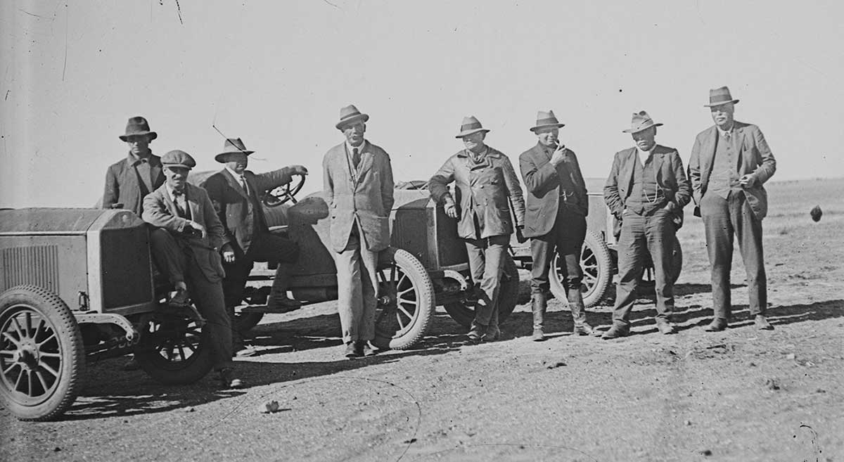 A group of men standing in front of two cars, 1923. The eight non-Aboriginal men all wear trousers, jackets, shirts, shoes, and hats. They stand in front of three 1920s open-wheeled cars. One man leans against the front of the middle car. Another stands at the side of that car, his left hand on its steering wheel and his left leg up on its running board. The man at the far left in the image sits on the front left tyre of the left side vehicle. He has his right leg drawn up on the car's suspension and rests his right elbow on the car's radiator. The ground around the men and cars is bare, with the exception of scattered small rocks. The horizon can be seen at the far right of the image; the rest of it is obscured by the men and cars.
