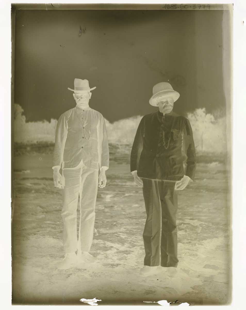 Glass plate negative of two men. They stand next to each other on open ground; the Aboriginal man is to the left in the image. He wears shoes, trousers, a shirt and a hat. The other man wears shoes, trousers, a shirt and a pith helmet. Both stand in stiff, formal poses with their hands by their sides. - click to view larger image