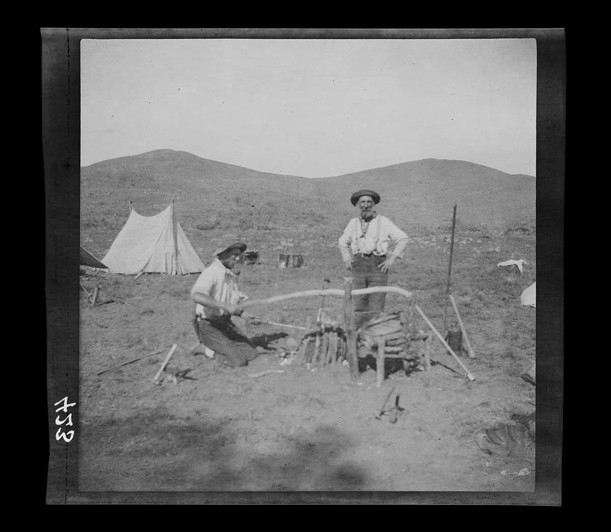 Two blacksmiths at work, Mount Davies camp, South Australia 1903. One kneels on the ground to the left, operating a small improvised furnace. The other stands to the right, hands on hips, facing the camera. Both are bearded and wear hats, light-coloured shirts and long trousers. A tent from the camp is behind them to the left. Two rounded hills are in the distance. - click to view larger image