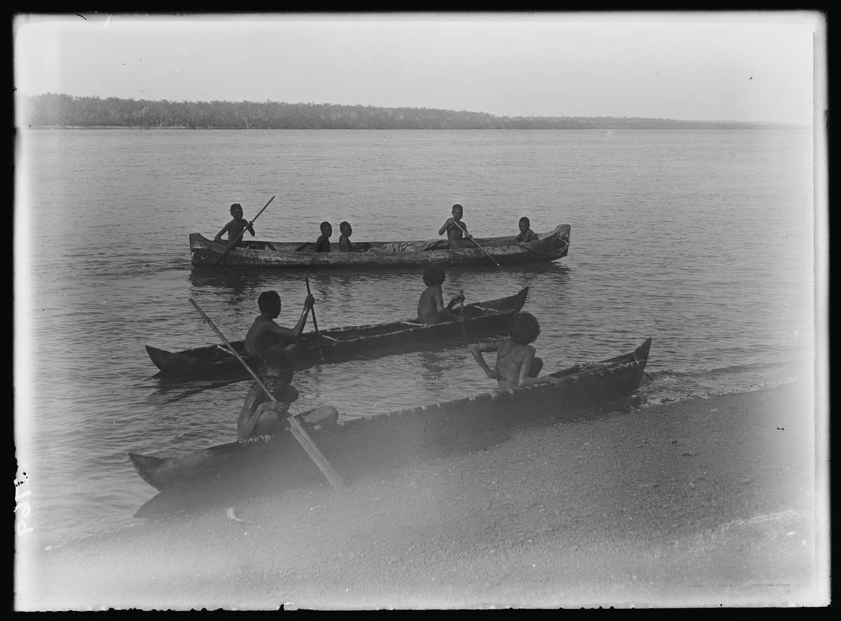 Nine Aboriginal boys in a dugout and two bark canoes. One canoe is at the water's edge in the foreground. The other two are just offshore, about parallel to the first canoe. Behind the canoes is open water and low hills on the distant opposite shoreline. The water's surface is slightly rippled. - click to view larger image