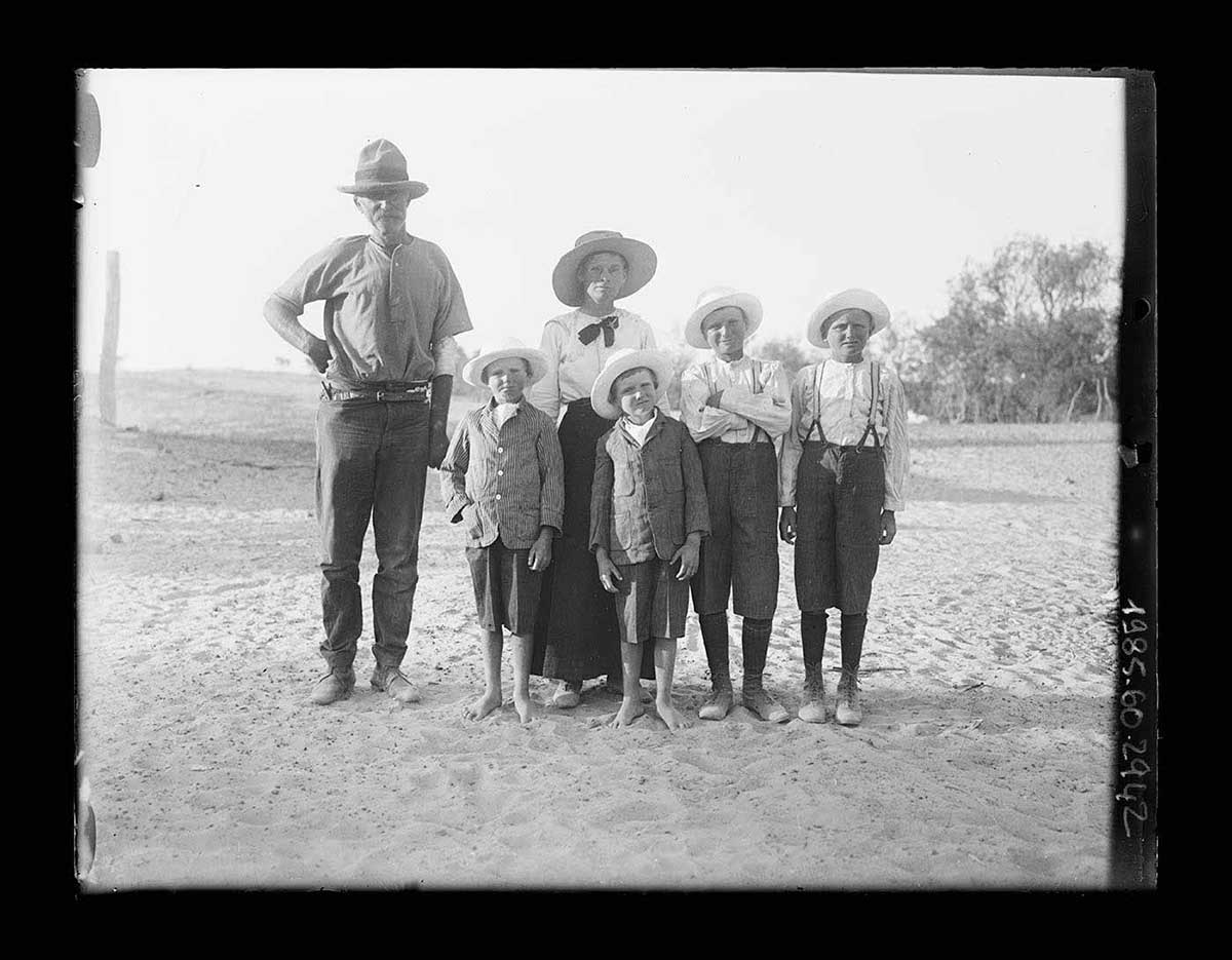 A family photo taken outdoors, Karrathunka Waterhole, South Australia 1919. The six members of the family stand in the centre of the image. The father stands to the left of the image, slightly apart from the others. The mother stands on his left. In front are four children, all boys. Two older boys dressed in long short pants, braces and hats stand to the right of the mother. Two younger boys dressed in jackets, short pants and hats stand in front of the mother. The group stands on dimpled sandy soil. - click to view larger image