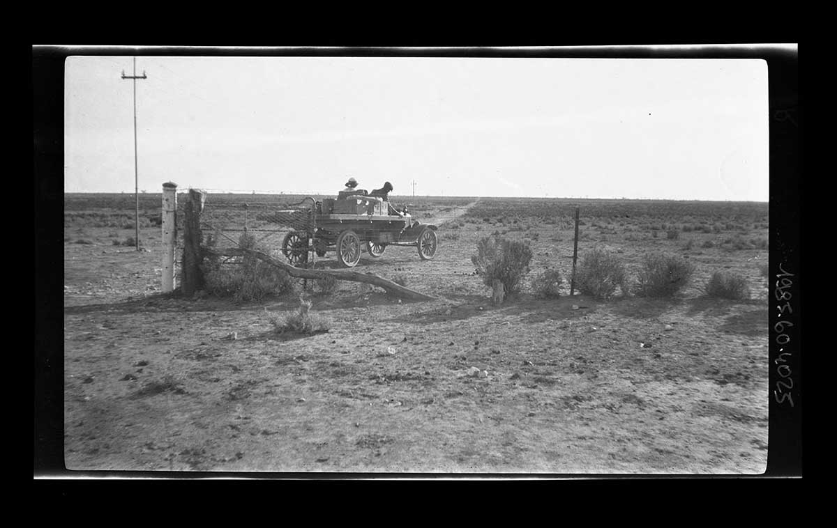 A T-Model Ford truck with two passengers, South Australia 1920. The truck has a flat tray body, upon which is stacked some equipment. The driver and a passenger are in the open cabin. The truck appears to have just driven through an open gate at the left side of the image. The gate is part of a fence that is between the truck and the camera. A single telegraph pole is at the far left of the image, rising up through the horizon line. - click to view larger image