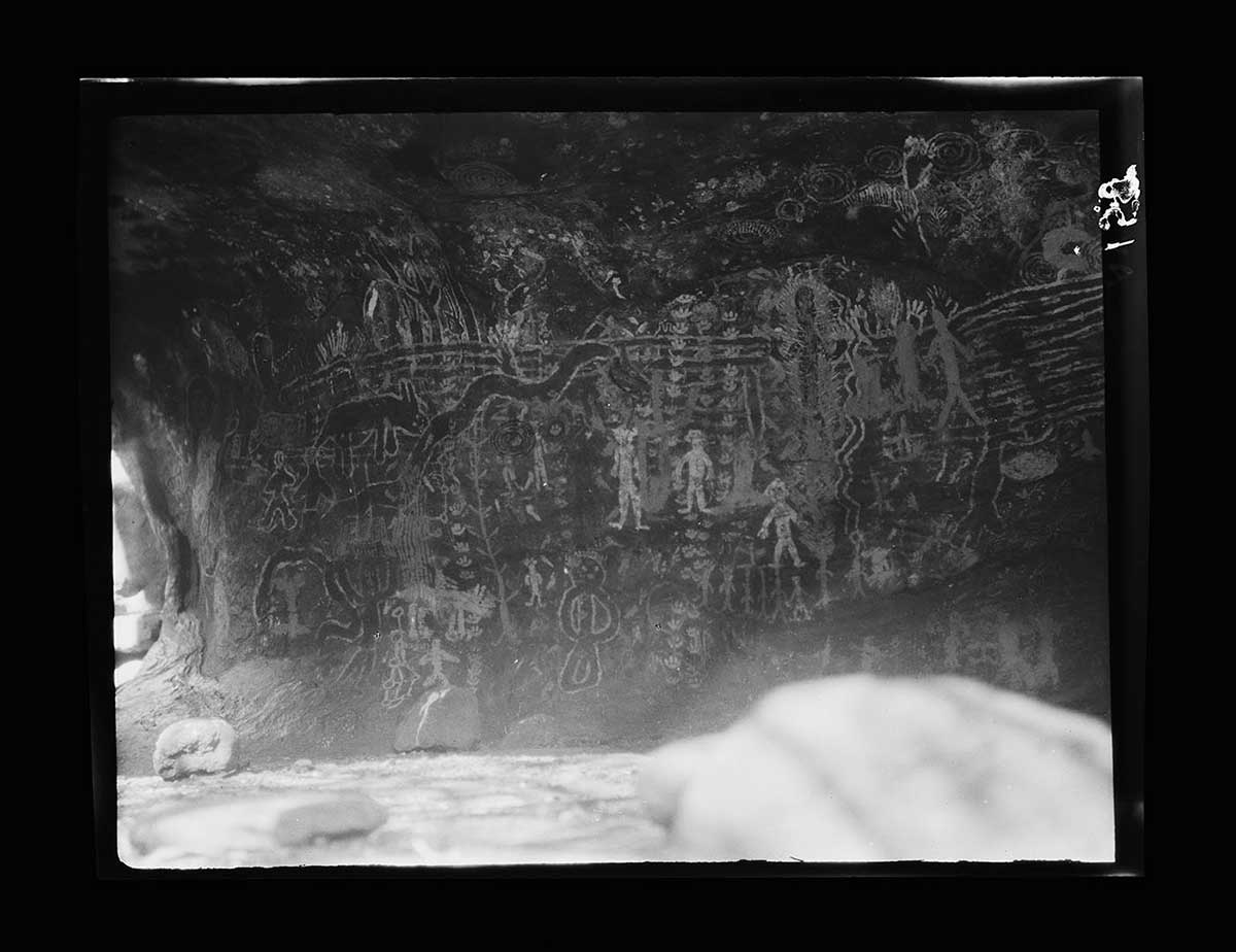 Rock paintings, southern side of Uluru, Northern Territory 1926. The paintings are on a rock wall that curves up gently from the ground. It is in shadow, suggesting that there is a rock overhang above. Much of the visible rock wall is covered in the paintings, which depict customary patterns, figures and animal totems. The paintings have been overlayed, creating a mult-layered effect. Some images are brighter than others, suggesting that they are more recent additions or perhaps in lighter colours. A few loose rocks are scattered in the foreground. One, in the front right hand corner if the image, is quite close to the camera and out of focus. - click to view larger image