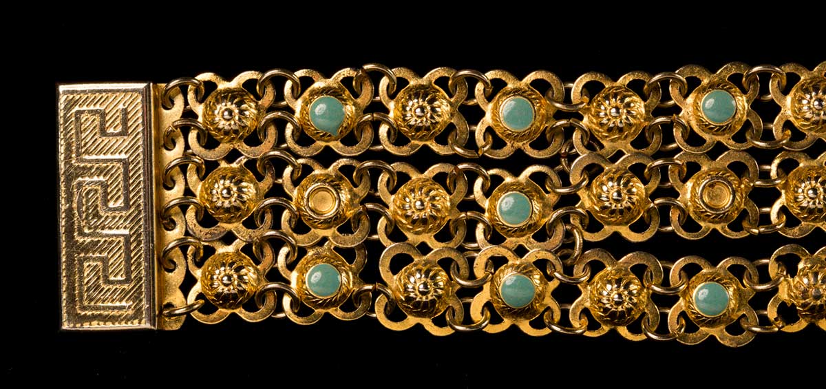 Photograph of details of a gold-coloured metal bracelet. The rectangular clasp of the bracelet has a Greek meander pattern. The bracelet consists of three chains of daisy-like units connected together by metal link chains. Every second line of daisy segments is decorated with a turquoise-coloured stone in the centre. A few of these turquoise stones are missing. - click to view larger image