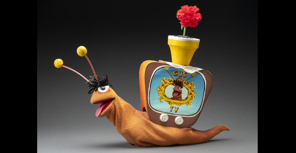 A puppet of a snail named Gus. Gus has an orange body, a tuft of black hair, orange and yellow antennas, brown eyes and a pink felt tongue. Attached to its back is a television box with two knobs, a screen featuring an image of Gus’s head surrounded by a gold frame, and printed text that reads ‘GUS/TV’. Attached to the top of the television is a yellow flowerpot with a red flower, sitting atop a doily.