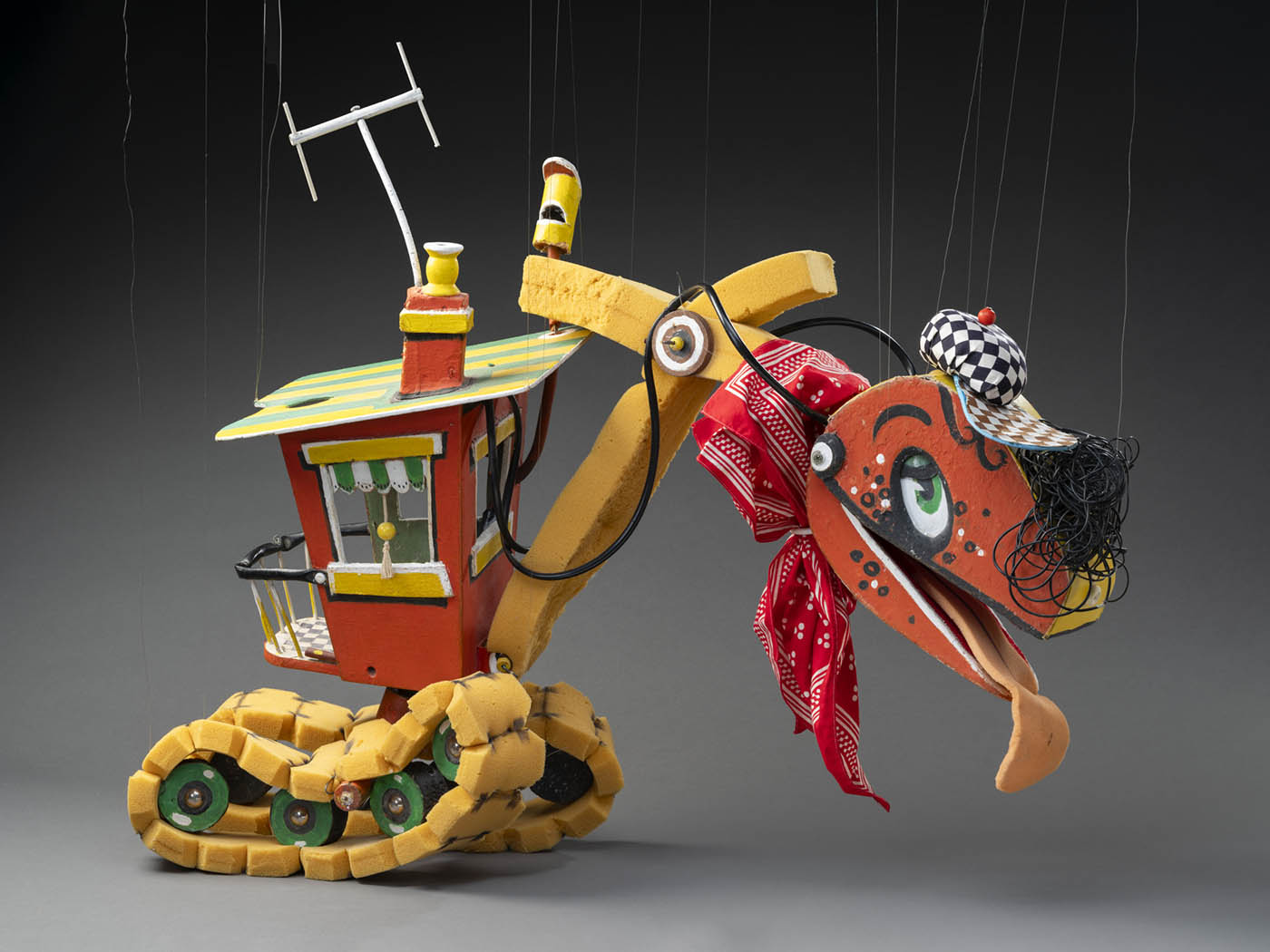 A puppet named Bill Steamshovel, in the form of an excavator. Bill Steamshovel consists of a red, yellow and green wooden control room connected to which are foam tread tyres and a foam neck. At the end of the neck is a red and yellow excavator head with a checkered hat and hair made from strands of black rubber. Tied around the neck is a red and white handkerchief.