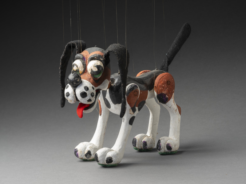 A photo of wooden puppet dog called Noodle. Noodle has a black, brown and white painted body, felt ears and green felt soles attached to its feet. Noodle has green painted eyes and a red felt tongue.