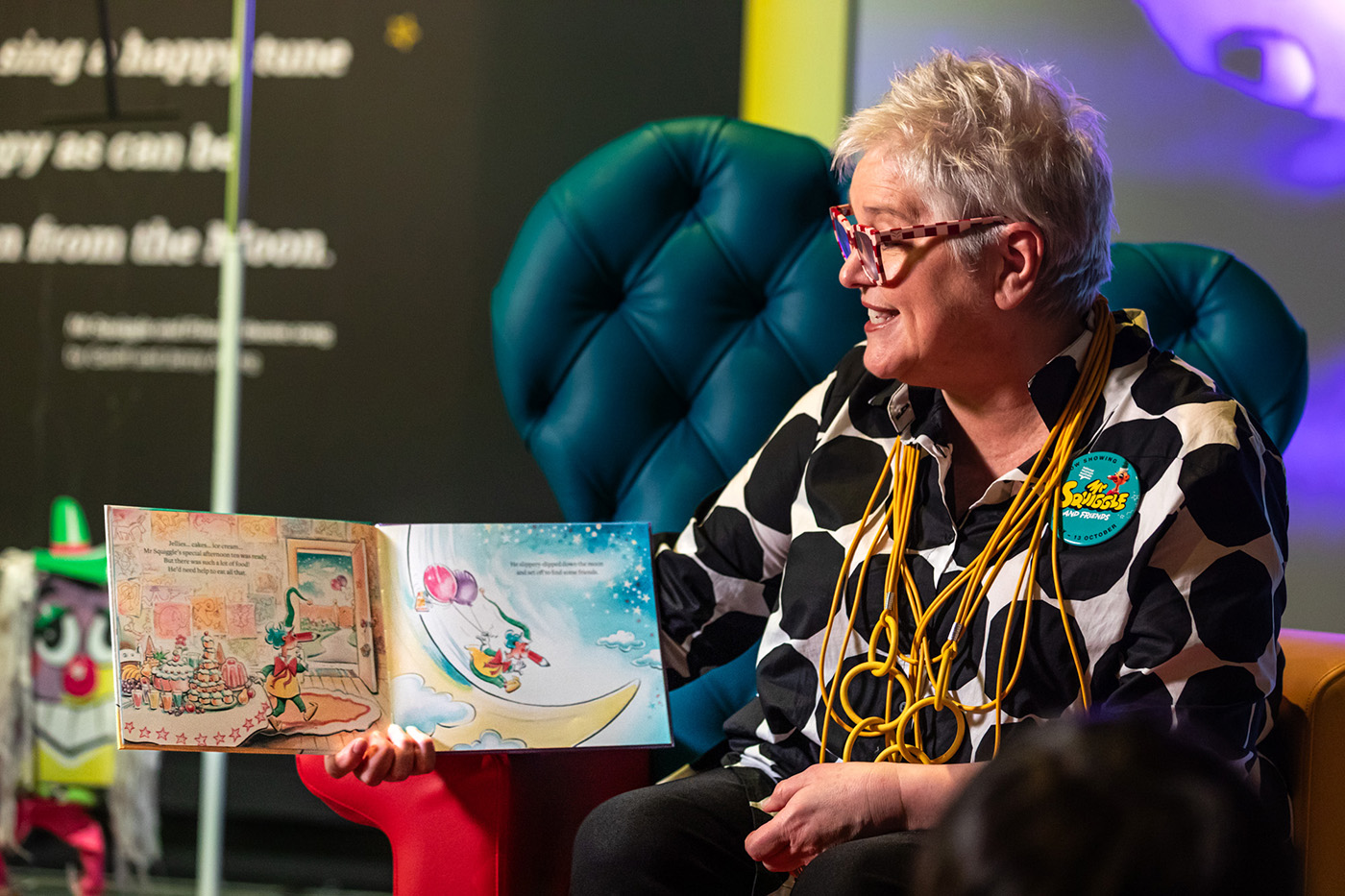 A woman sits on a high-backed chair, reading from a children’s book, held open on pages showing brightly coloured images of Mr Squiggle. 