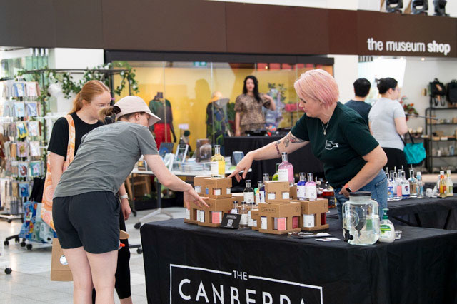 Two women look at a display of small bottles on a table, with another woman standing behind the table, pointing at a product. - click to view larger image