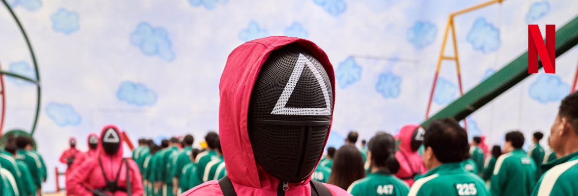 A man in a hooded pink tracksuit, with a black face mask with a white triangle symbol on the forehead, stands amid a field of people, some wearing green tracksuits, unmasked, and others wearing black face masks and pink tracksuits.