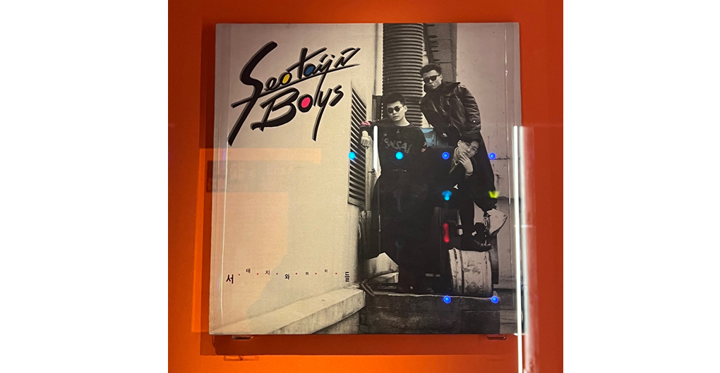 This album cover for Seo Taiji and Boys' self-titled debut album 'Seo Taiji and Boys' (1992) is in a black and white, with a gritty 90s urban look. On the cover are the three members of Seo Taiji and Boys, posing in a narrow industrial alleyway. They are casually leaning against ductwork, conveying a 'Street' attitude. They are dressed in dark, edgy 90s fashion, with sunglasses and leather jackets. The name of the group, Seo Taiji and Boys, is displayed in a graffiti-like scrawl in the upper left of the record cover, with three letters marked out in yellow, blue and red. Additional Korean text is at the bottom spelling out the name of the group.