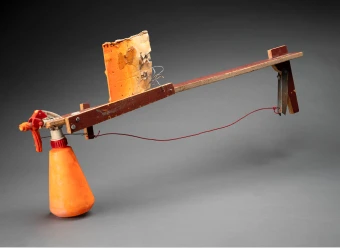 Four pieces of structural plywood screwed together in narrow strips to make a gun-like frame, with a wire connected to the trigger mechanism of a plastic spray bottle which is operated by a tinplate trigger. The bottle contained orange paint and is held in place with two small wires twisted through the top of the bottle and the plywood frame. It also has a handle and a simple cardboard target painted orange is connected by wire to a movable plywood arm screwed to the frame near the plastic bottle.