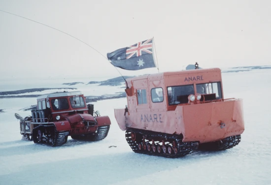 Two tracked vehicles driving across an icy Antarctic landscape.