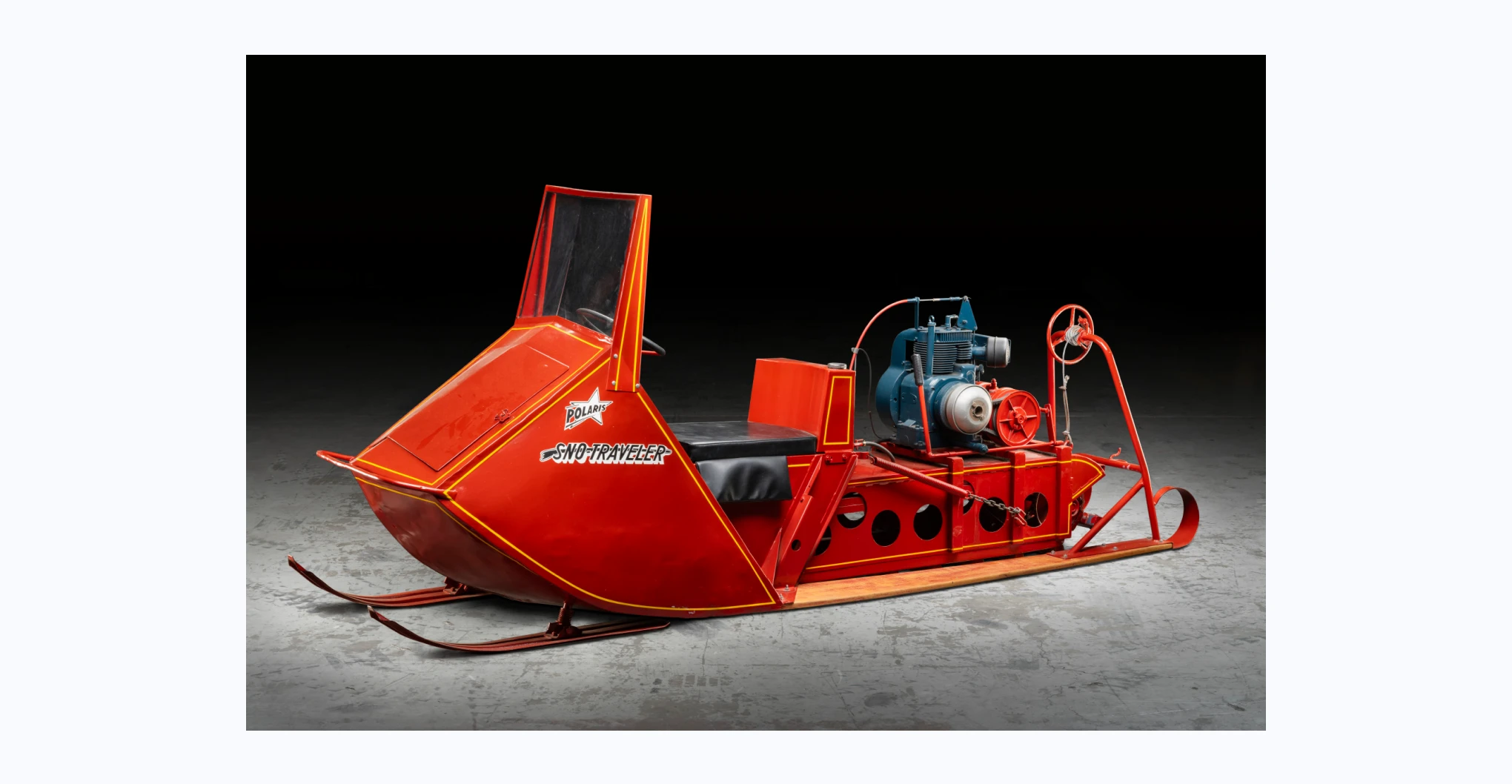 An oversnow vehicle, painted red with yellow pin stripes. The vehicle has metal sledge tracks at the front and a caterpillar track and wooden skis at the back. There is a latched door in front of the windscreen. The open driver’s cab has a half steering wheel, brake and throttle levers and an ignition switch on the right of the dashboard. The driver's seat is upholstered in black leather and the seat back is the fuel container. The engine is mounted behind the fuel container and a winch is mounted on the rear of the wooden skis. Logo with text painted in white and black on the right side reads ‘POLARIS / SNO-TRAVELER’. 