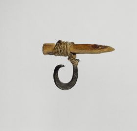 Fishhook made of mother-of-pearl that is supported at the upper end of the shank where strings made of various plant fibres are attached and tied to a small piece of pale, light wood.