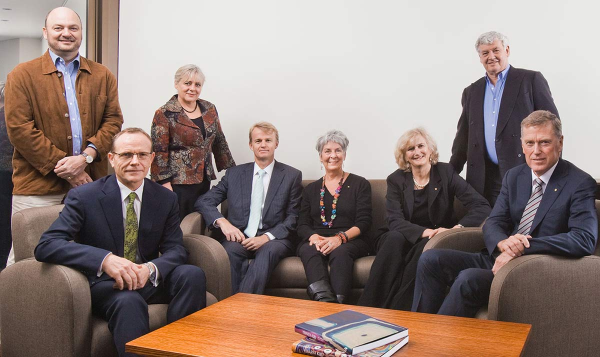 Group portrait showing three men and two women seated on sofas, with another two men and a woman standing behind. A low honey-coloured timber table, with two books on centre top, is in the foreground.