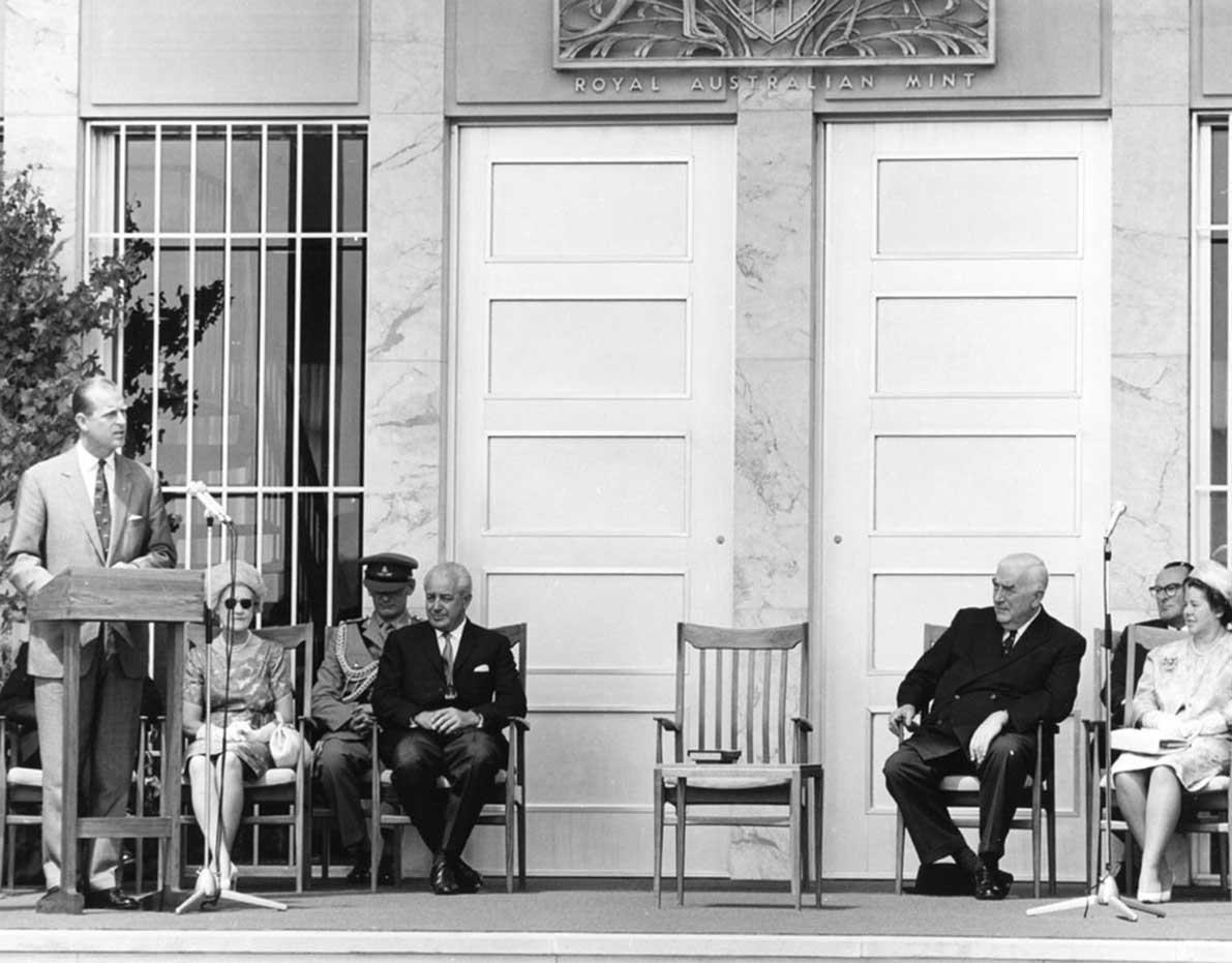 Prince Phillip speaking at a lectern with Holt and Menzies seated. Other dignitaries and their wives are also present. Behind them is the main door of the Mint. 