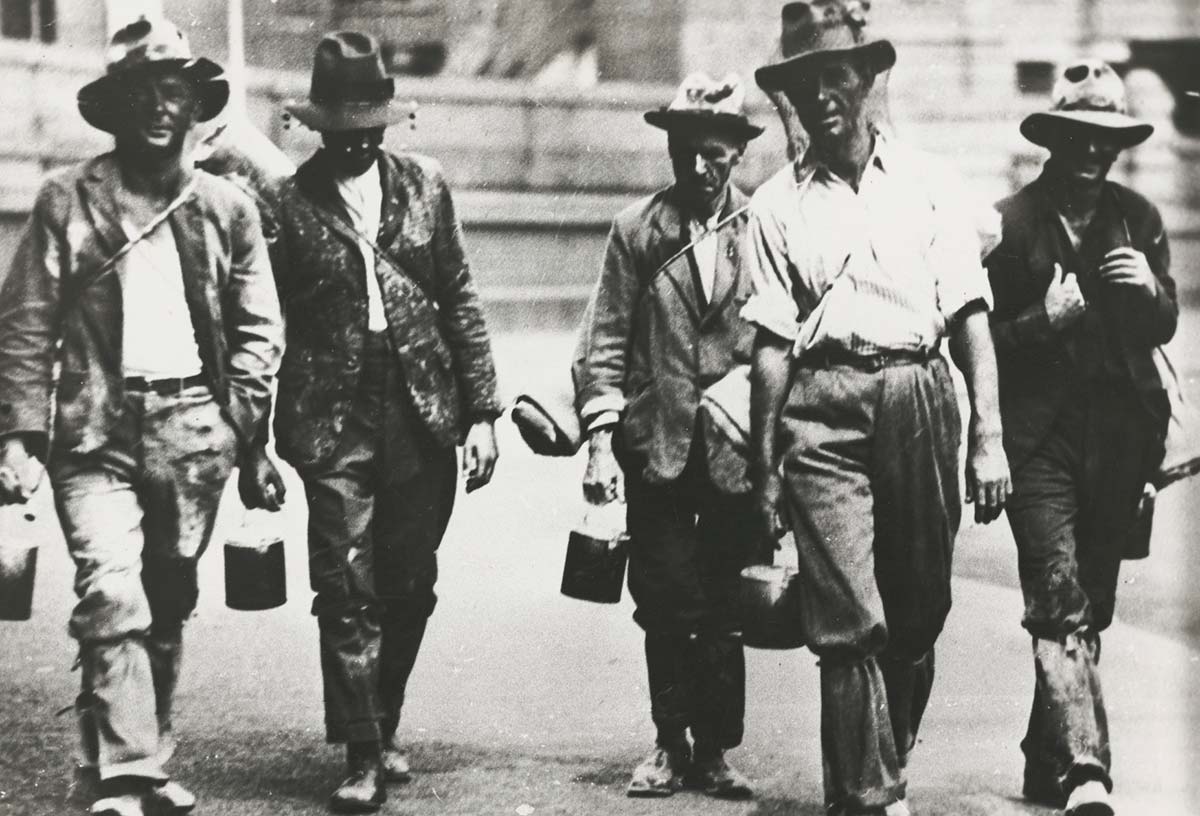 Five men walking towards the camera dressed in shabby clothes. Most are carrying swags and bill cans