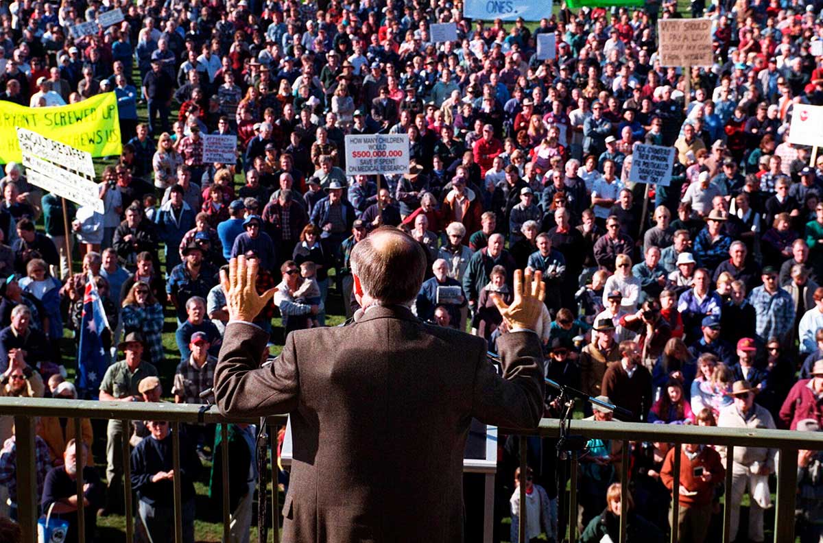 Photo taken from behind Prime Minister Howard who is wearing a bullet-proof vest under his jacket. Facing Howard is a crowd, some of whom are holding placards.