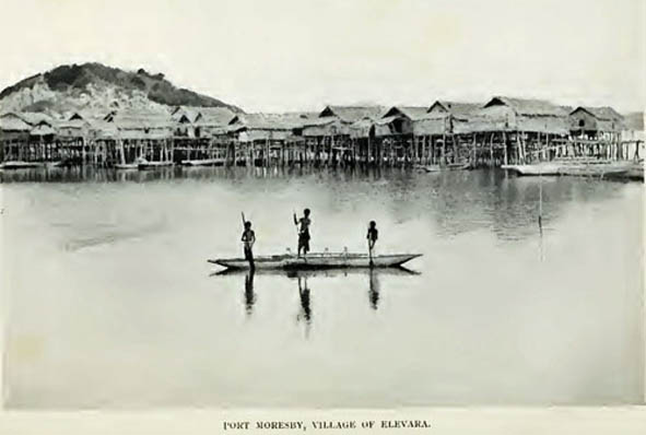 Black and white photo of canoes on a body of water with a village on stilts in the background. - click to view larger image