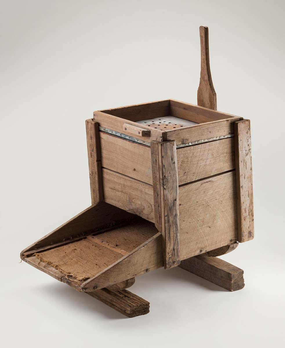 Studio photo of a simple contraption made of timber. It appears to be essentially a box with a tray extending from one side, and a handle from the other. On the top surface of the box is a metal sheet punctuated with holes. - click to view larger image