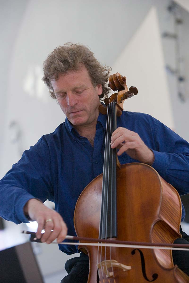 A man sits on a bench holding a wooden stringed instrument. The neck rests on his right shoulder and he draws a bow across it with is left hand. - click to view larger image