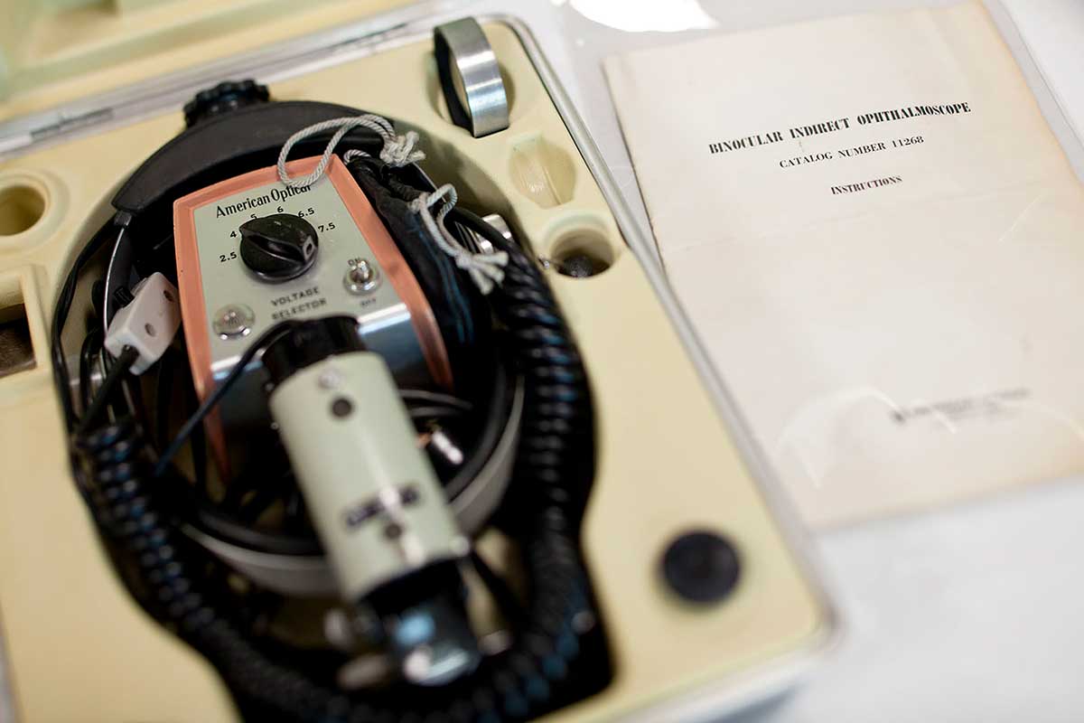 An 'American Optical' brand small handheld othalmoscope stored in a hard, grey plastic case. The case also contains a power adapter and cables and sits alongside a paper instruction manual for a 'BINOCULAR INDIRECT OPTHALMOSCOPE Catalog number 11268'. - click to view larger image