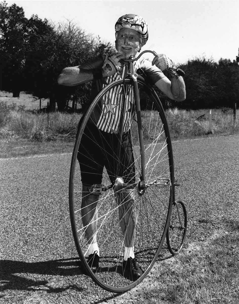 Black and white photograph of a man standing beside a penny-farthing bicycle. The man, who wears a helmet and has zinc cream across his cheeks, rests his head on the handlebars. He wears cycling gloves, shorts and a shirt with stars and stripes at the front. - click to view larger image