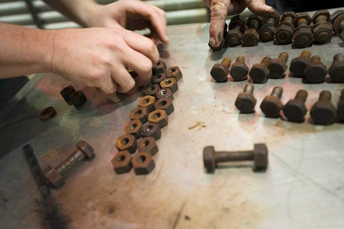 Detail shot showing two hands at the right, arranging two rows of rusted nuts. Another hand extends from the top of the image, to rest on a row of bolts. - click to view larger image