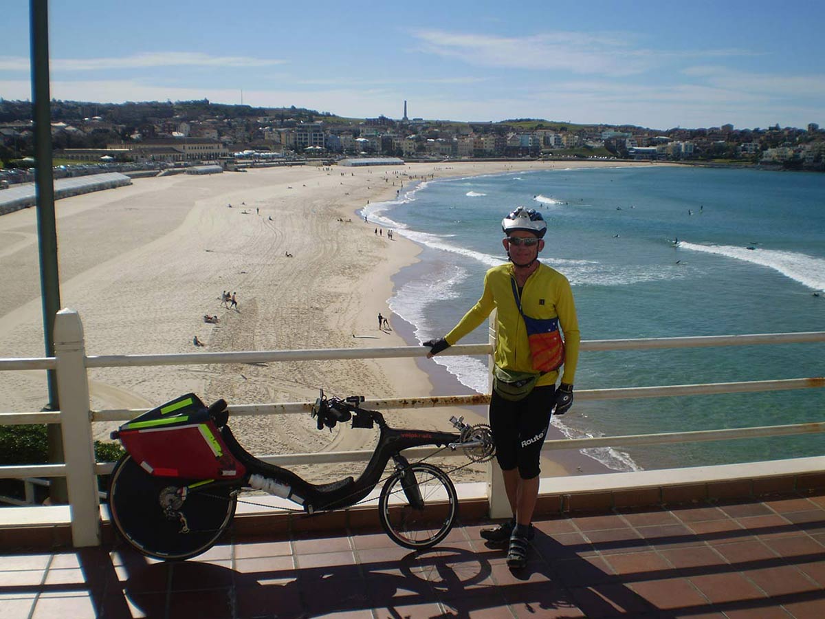 Colour photograph of a cyclist standing beside a recumbent bicycle. Bondi Beach is visible in the background. - click to view larger image