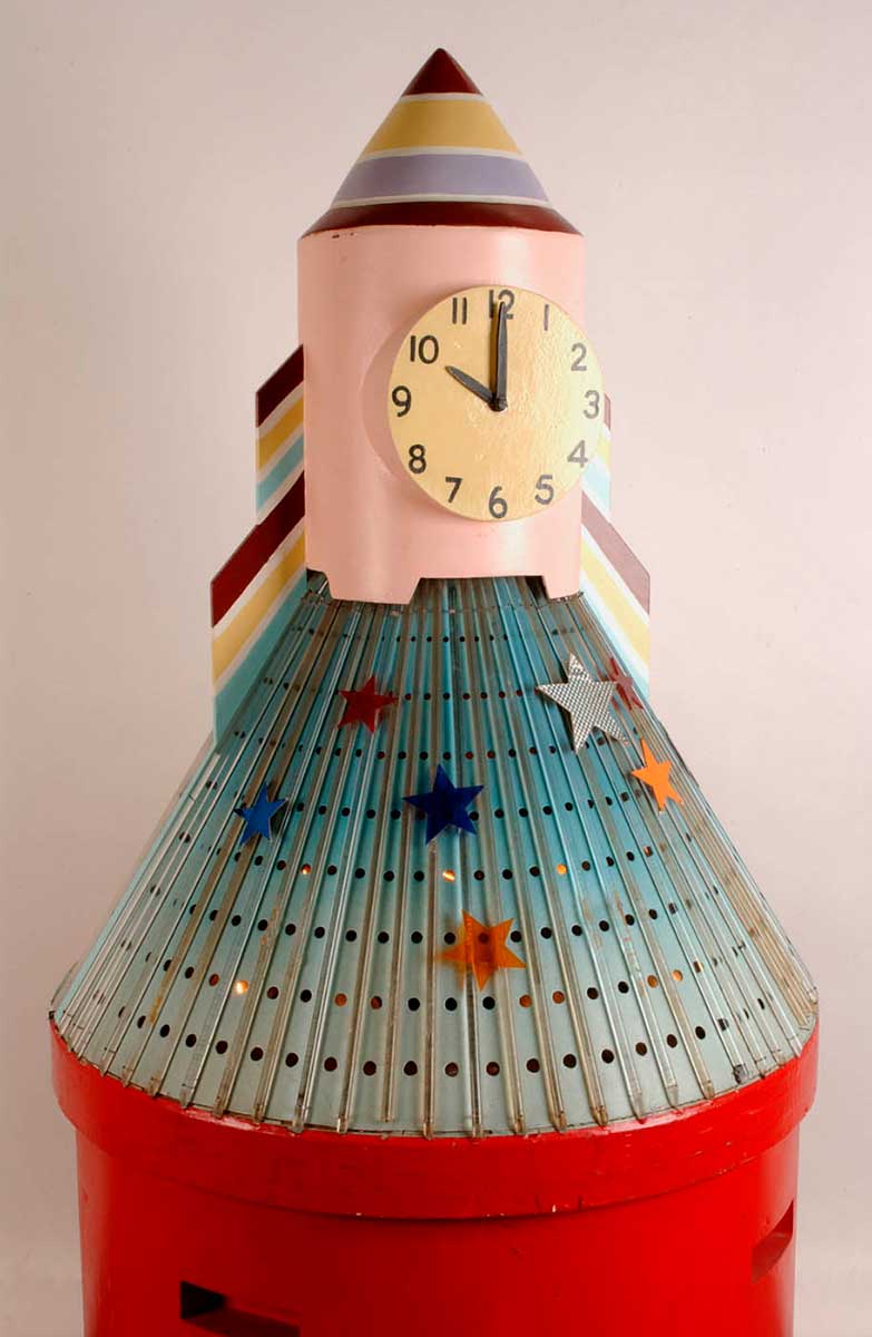 A large clock in the shape of a rocket with a red cylindrical wooden base with a light blue perforated metal skirt covered in vertical plastic rods, a light pink wooden trunk and painted striped cone top. - click to view larger image