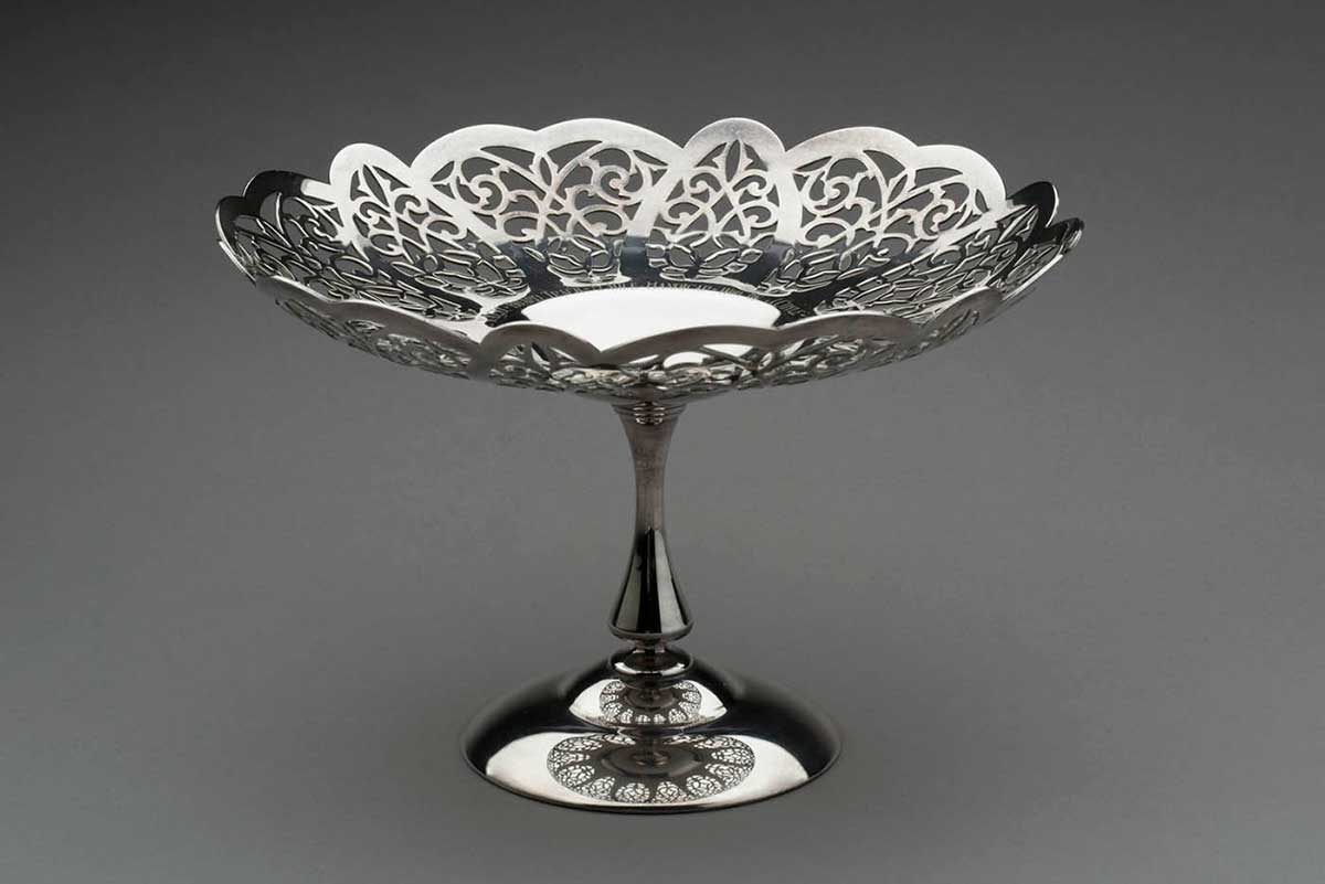A silver-plated bowl shaped trophy, with a scrollwork design at the top and a thin vertical stand and round base. - click to view larger image