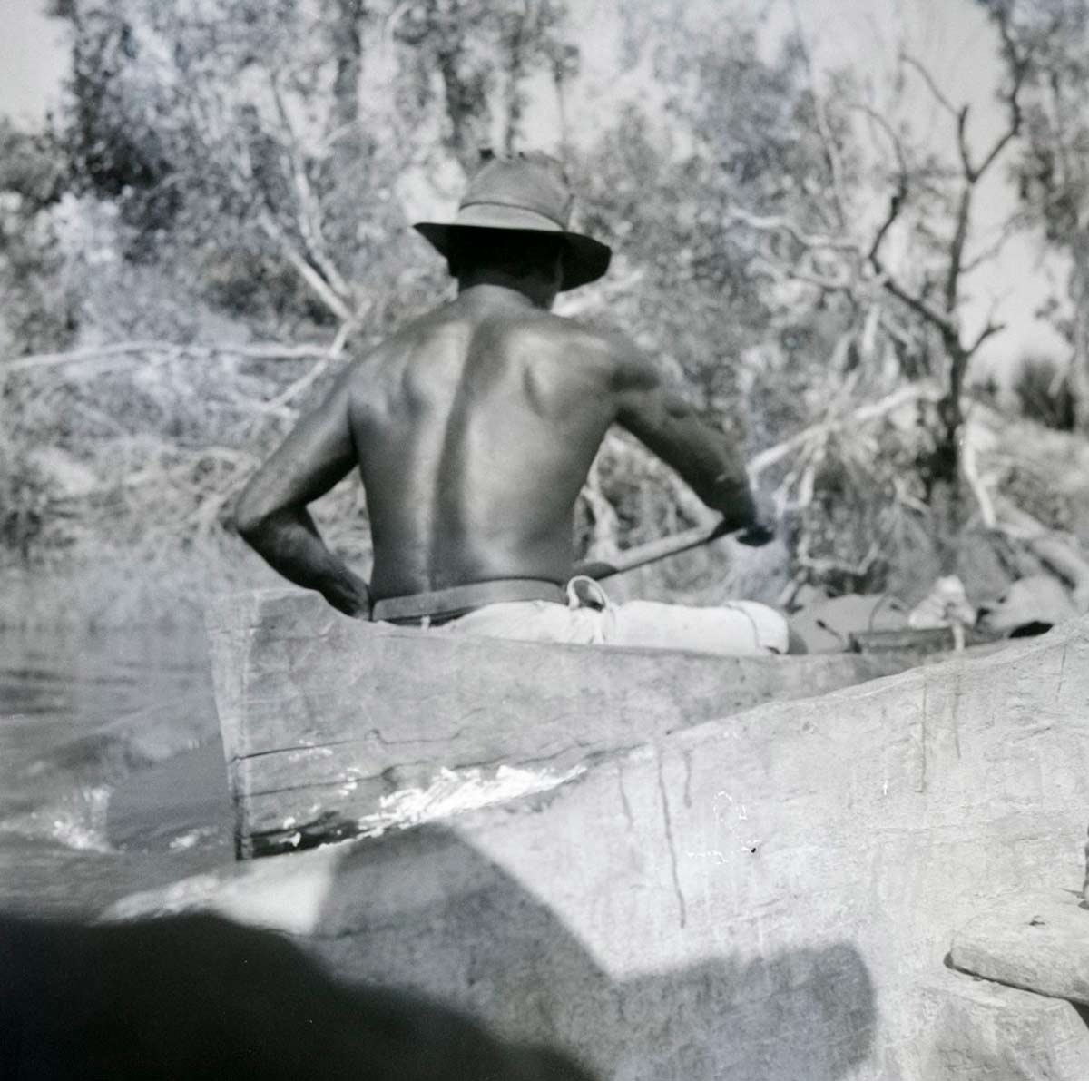 A black and white photographic negative that depicts an Aboriginal man wearing a hat paddling a canoe with his back to the camera. - click to view larger image