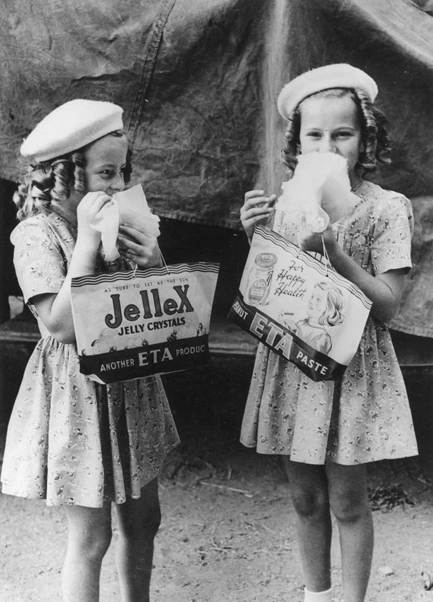 Two young girls eat a treat while holding show bags. One reads 'Jellex Jelly Crystals'; the other 'Peanut ETA Paste'. - click to view larger image