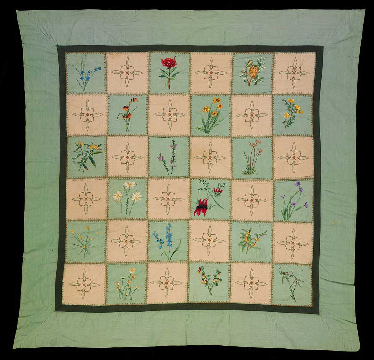 Fabric quilt with green and cream patchwork squares embroidered by hand with Australian wildflower designs, and a recurring passionfruit vine flower design.