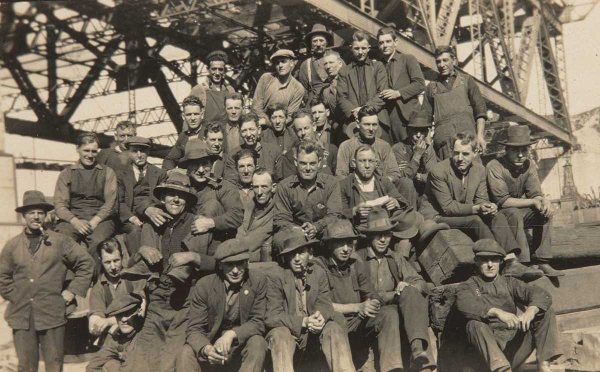 A group photograph of workers sitting under a large steel construction. Several smoke pipes and most wear overalls and hats. - click to view larger image