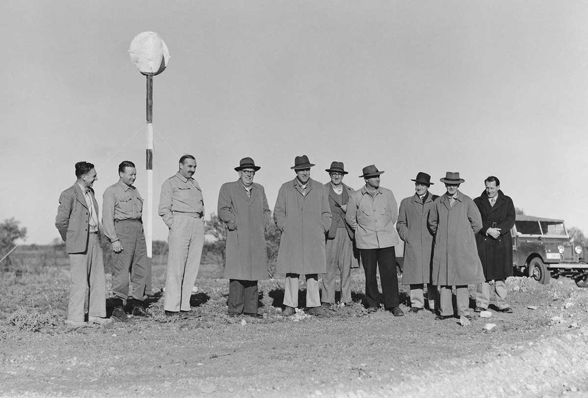 A group of 10 men pose on a dirt track with a jeep in the background.