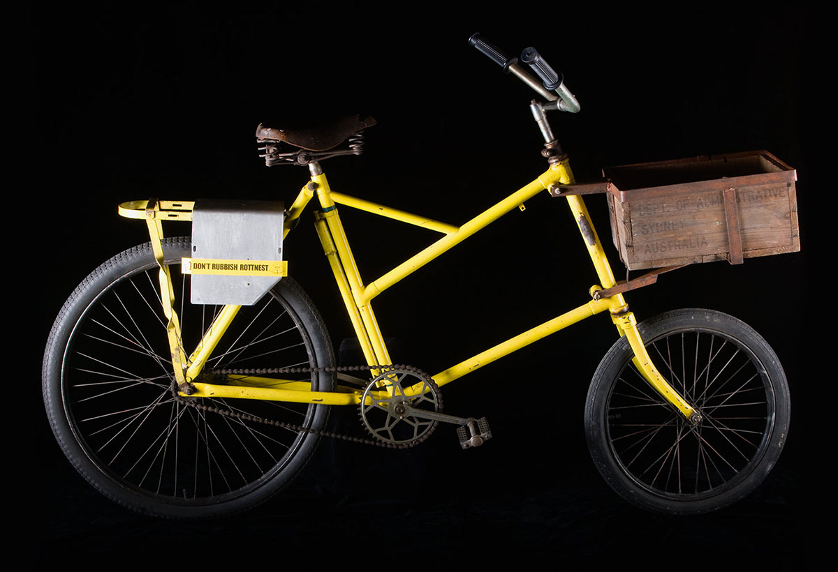 A bright yellow bicycle with a wooden box fixed to the front and a sign affixed to the rack that reads ‘DON’T RUBBISH ROTTNEST’. - click to view larger image
