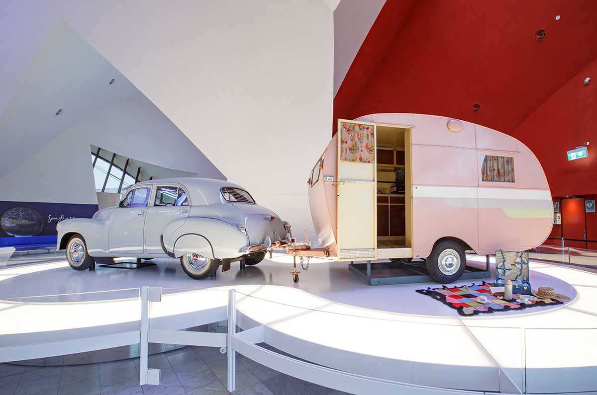 Display featuring a vintage car attached to a vintage pink caravan. A picnic set is laid out on a blanket in front of the caravan.