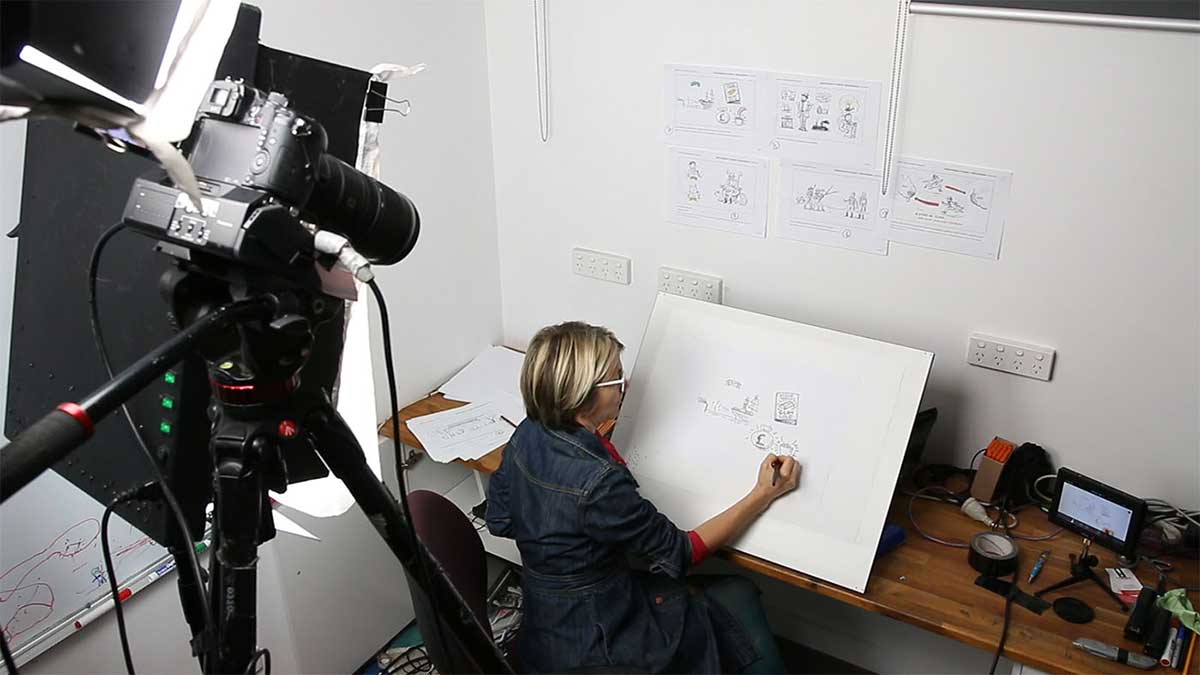 A woman seated in front of an easel. being filmed by a video camera. - click to view larger image
