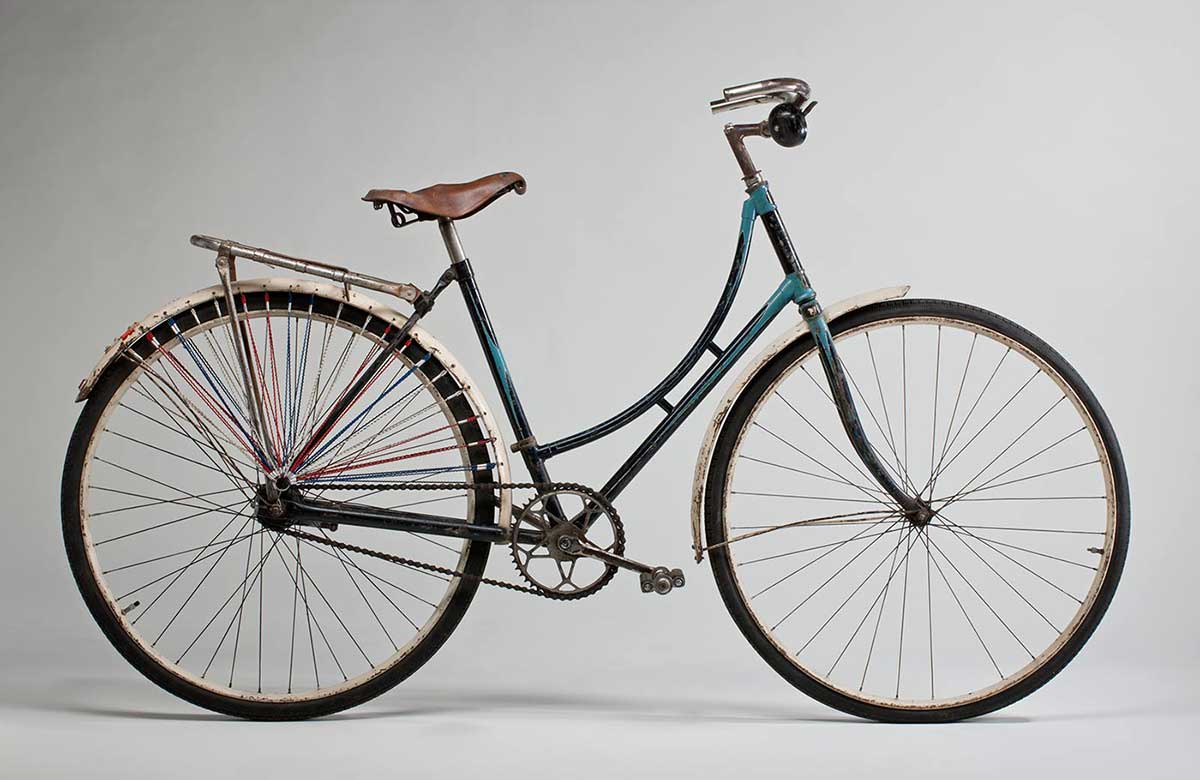 Ladies refurbished 'Roadmaster' bicycle. The frame is handpainted in turquoise and navy blue enamel paint, and the mudguards are white. It has a brown leather seat and a silver rack attached to the rear end. Both of the wheels are from a Malvern Star. The rear wheel has lengths of red, white and blue fine cord attached through the mudguard and gathering just above the centre of the wheel; on both sides of the rear wheel. Small reflector on rear wheel guard. Large black bell on right side of the handlebar. - click to view larger image