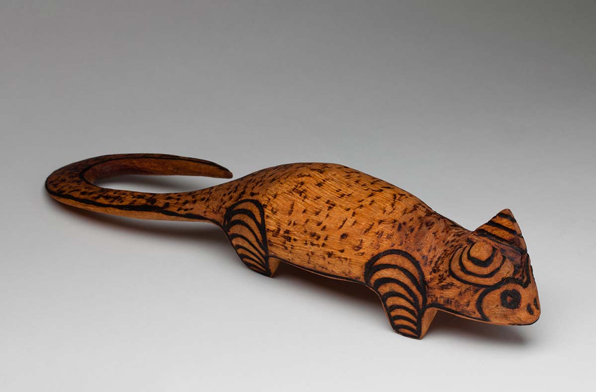 Small black and brown wooden cat sculpture standing on four short legs with the top exterior covered in burnt pokerwork designs.The triangular shaped head has burnt markings around the eyes, nostrils and mouth while the two ears are pointed and decorated with concentric semi-circles. The side of the head, neck, body and long hooked curled tail are decorated with a lightly shaded speckled pattern and the four legs are covered with parallel, horizontal curved lines. - click to view larger image