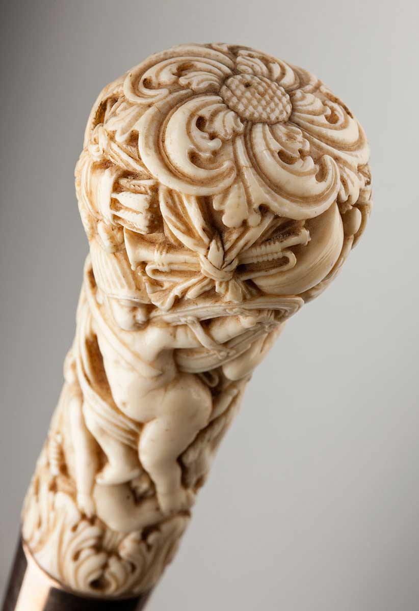 Studio colour photo of ivory grip, carved with a range of motifs, including what looks like a baby. - click to view larger image