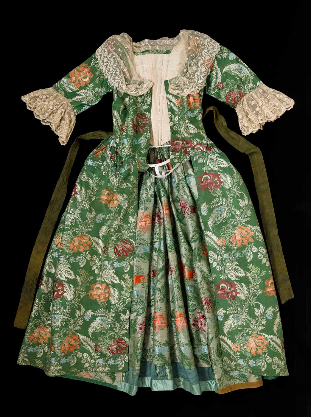 The front of a green and floral dress with lace trim on the sleeves and collar with a long green sash like belt stitched at the waist. - click to view larger image