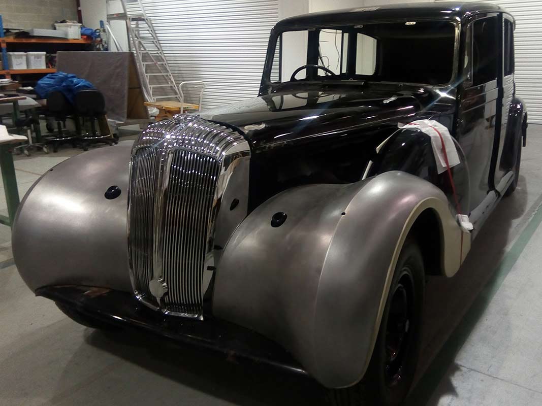 An old, sedan-style car with new black paint on the body. The front wheel hubs are dull grey and unpainted. The car has no headlights or glass in the windows.