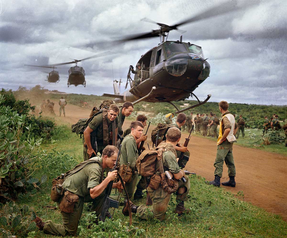 Seven young soldiers crouch down while a helicopter lands next to them on a dirt road in open countryside.